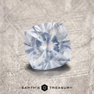 A 1.32-Carat Pale Blue Montana Sapphire with a cushion cut sits on a textured metallic surface, with "EARTH'S TREASURY" displayed below.