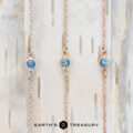 A trio of pendant necklaces, each graced with alluring blue gemstones on chains of silver, gold, and rose gold, set against a wooden backdrop. A touch from the Lumi collection infuses the ensemble with the charm of Earth's Treasury.