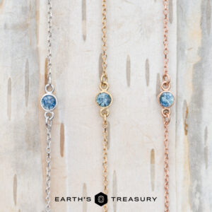 A trio of pendant necklaces, each graced with alluring blue gemstones on chains of silver, gold, and rose gold, set against a wooden backdrop. A touch from the Lumi collection infuses the ensemble with the charm of Earth's Treasury.
