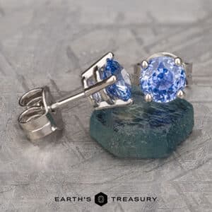 A pair of Medium Blue Sapphire Earrings, 5.5mm (Heated) with silver settings are displayed, with one earring standing upright and the other lying on its side on a textured surface. The text "EARTH'S TREASURY" and a hexagon logo are visible at the bottom of the image.