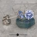 A pair of elegant Light Blue Sapphire Earrings, 6.0mm (Heated), featuring round lavender-blue gemstones set in shiny metal mounts. One earring is laid on a teal stone, showcasing its side profile, while the other stands upright. The background has a textured, grayish surface.