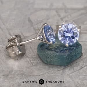 A pair of elegant Light Blue Sapphire Earrings, 6.0mm (Heated), featuring round lavender-blue gemstones set in shiny metal mounts. One earring is laid on a teal stone, showcasing its side profile, while the other stands upright. The background has a textured, grayish surface.
