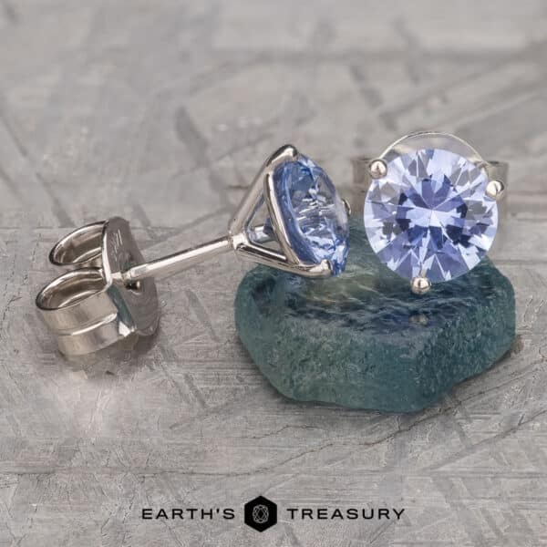 A pair of elegant Light Blue Sapphire Earrings, 6.0mm (Heated), featuring round lavender-blue gemstones set in shiny metal mounts. One earring is laid on a teal stone, showcasing its side profile, while the other stands upright. The background has a textured, grayish surface.