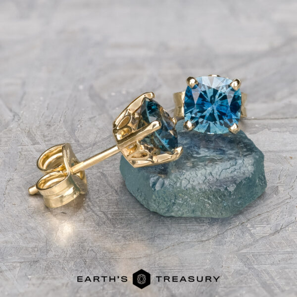 The enchanting Montana Sapphire Earrings, product code EA-MS-418, feature two cushion-cut stones measuring 5.0mm each, beautifully set in gold. One of the earrings rests elegantly on a textured blue surface with "Earth's Treasury" inscribed below.