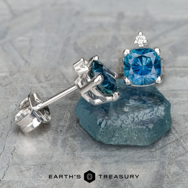 Displayed are two blue Montana Sapphire earrings with silver studs, with one resting on a matching cushion-cut stone. Set against a textured grey background, the text below reads: "EARTH'S TREASURY." These heated gems, known as EA-MS-431, boast an exquisite natural allure perfect for any occasion.