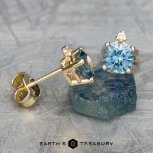 Gold earrings adorned with aquamarine gemstones and a heated cushion-cut green-blue stone are elegantly showcased on a textured surface, echoing the style of the EA-MS-432 earrings.