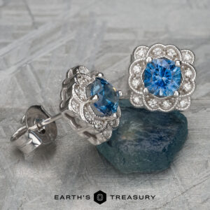 The EA-MS-511, silver earrings featuring cushion-cut blue gemstones and diamond accents, are showcased on a textured gray surface with a raw Montana Sapphire in the background.