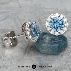 The EA-MS-516 earrings, featuring two exquisite Montana Sapphires each encircled by a halo of clear stones, are elegantly displayed on a textured surface, with one nestled atop a rough teal stone.