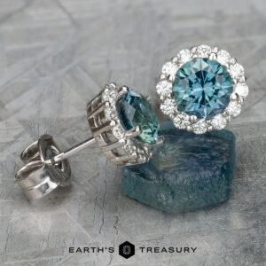 The EA-MS-519 earrings are a stunning pair of halo earrings showcasing large Montana sapphires, whose teal brilliance is elegantly complemented by smaller diamonds on a textured surface.