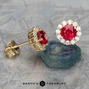 The EA-DB-504 earrings, featuring 5.0mm heated red stones surrounded by sparkling diamonds, are elegantly displayed on a textured surface.
