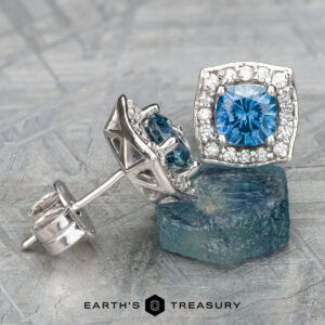 The EA-MS-520 earrings, featuring a stunning combination of blue topaz and diamonds set in a halo design, are elegantly showcased on a rough blue topaz stone.