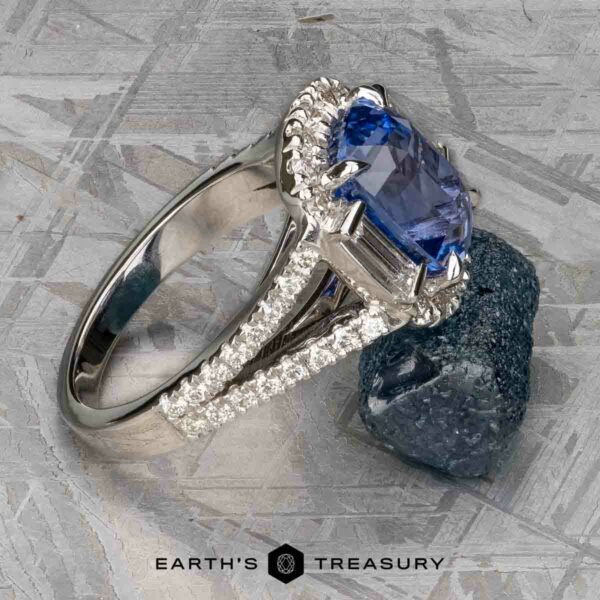 Vintage Halo Ring with 5.09-Carat Ceylon Sapphire in 18k white g Next to a rough blue mineral, an exquisite 18k white gold vintage halo ring features a striking 5.09-carat Ceylon Sapphire, accented with smaller clear stones on the band. The text "Earth's Treasury" at the bottom celebrates the elegance of this masterpiece.