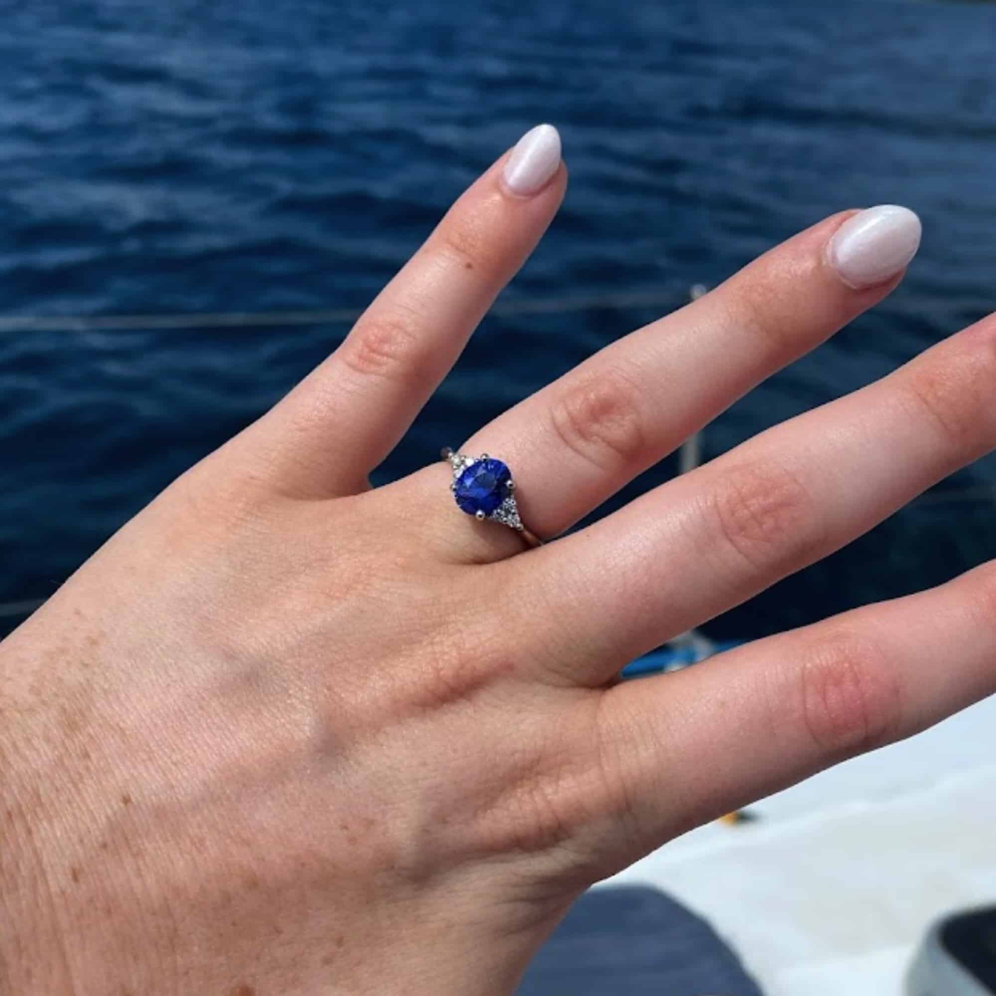 An engagement photo from a customer review featuring a "Kacie" ring with a royal blue oval sapphire, on a well-manicured hand against ocean waves