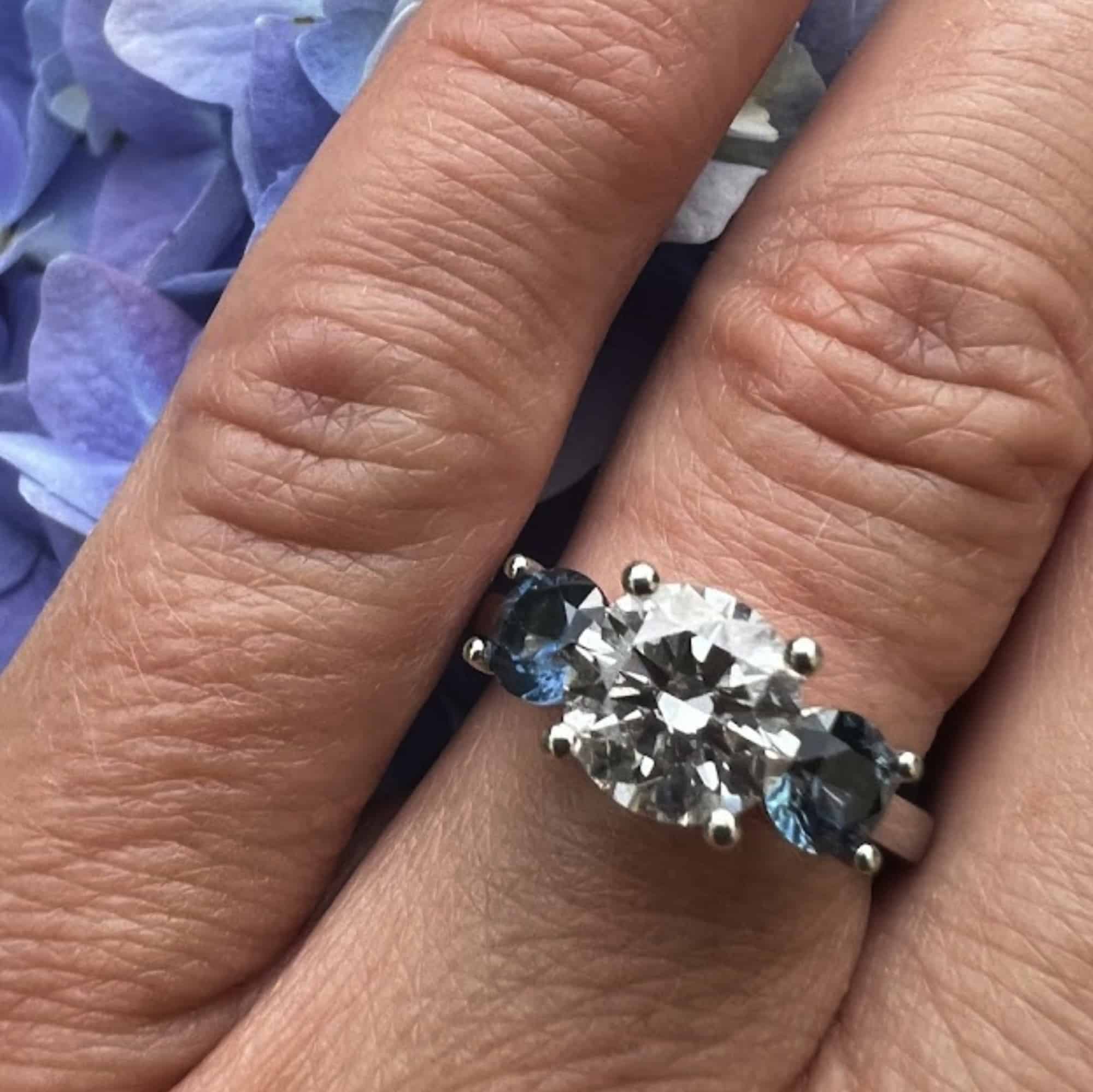 A photo from a customer review featuring a custom ring with heirloom diamond and Montana sapphire side stones, on a hand against light violet hydrangeas