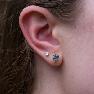 Close-up of a person's ear with two earrings: a small round stud and a larger cushion Montana Sapphire earring, featuring a heated sapphire gemstone (_DSC0507-1200-BW) in the earlobe.
