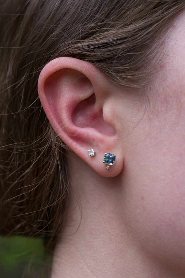 Accented Montana Sapphire Earrings Close-up of a person's ear with two earrings: a small round stud and a larger cushion Montana Sapphire earring, featuring a heated sapphire gemstone (_DSC0507-1200-BW) in the earlobe.