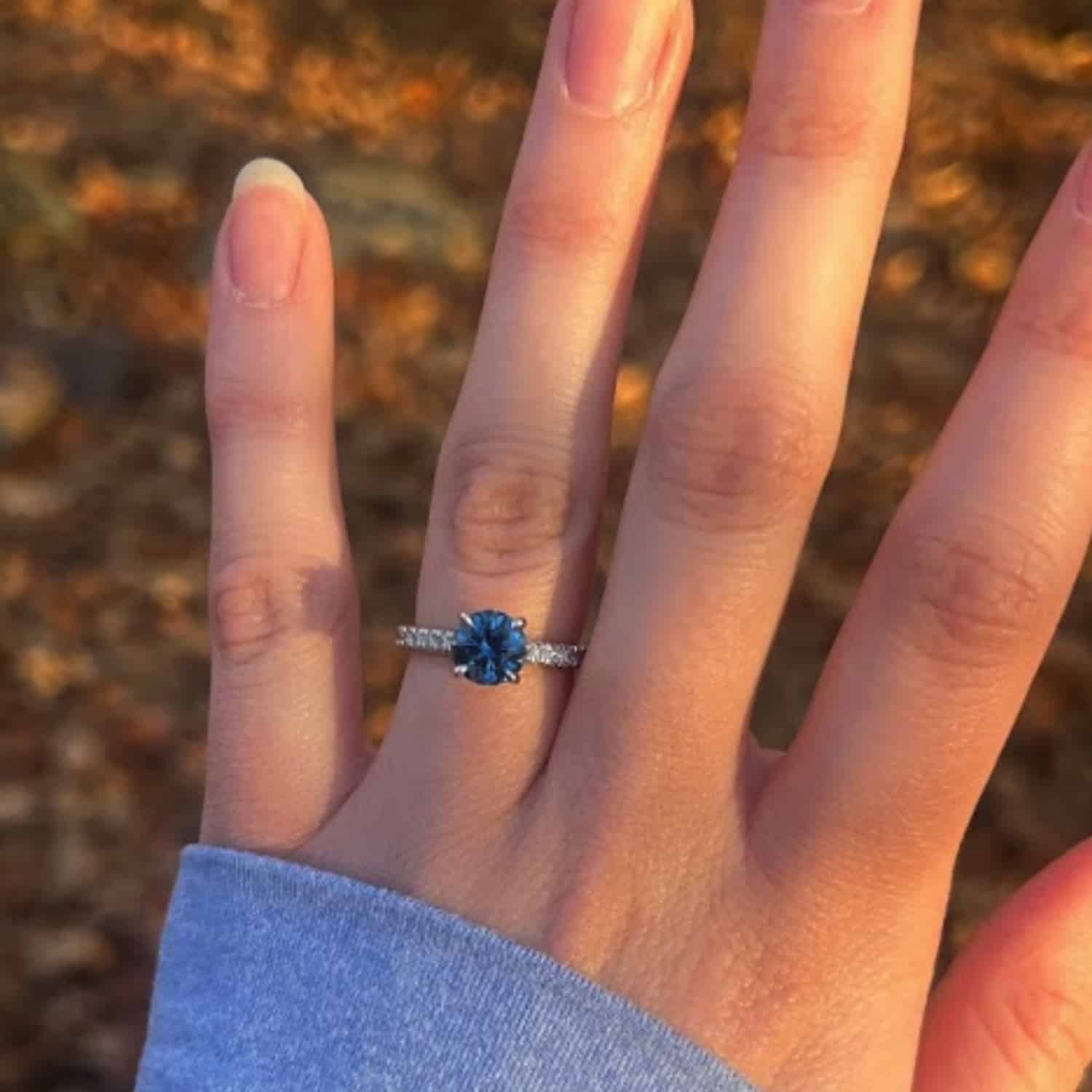 An engagement photo from a customer review, featuring a "Marina" ring with blue Montana sapphire on an elegant hand against a backdrop of fall leaves.
