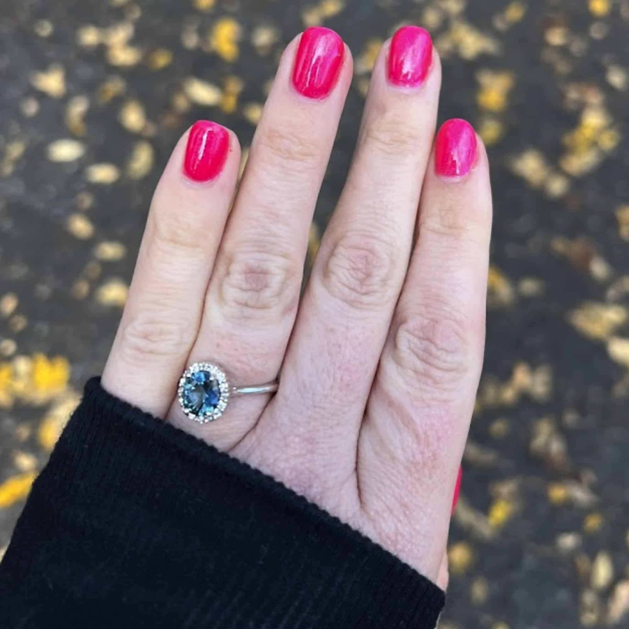 A photo from a customer review showing an "anne" ring with large teal Montana sapphire on a hand with hot pink nails against a ground scattered with fall leaves.