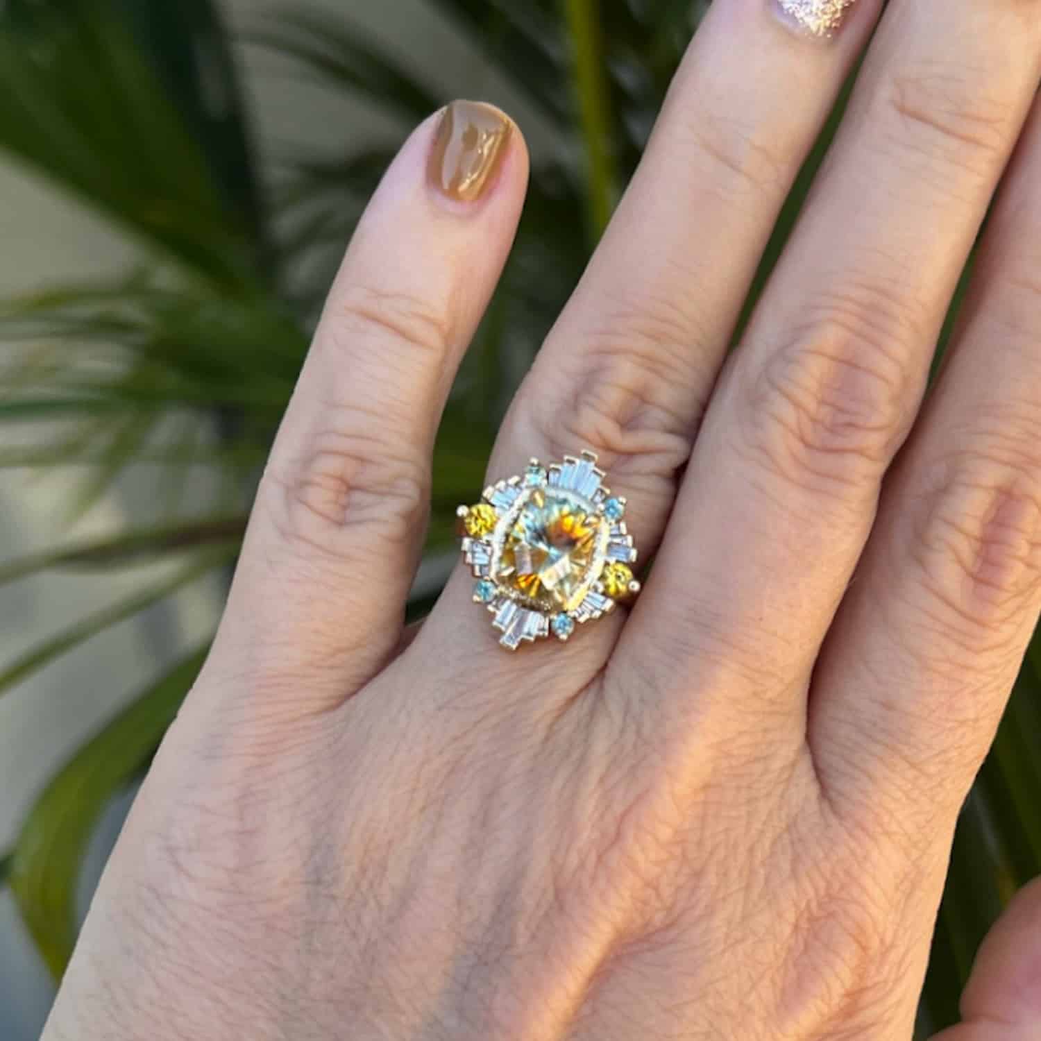 A photo from a customer review featuring a custom halo ring with a large particolored Montana sapphire, on a well manicured hand against a green houseplant.