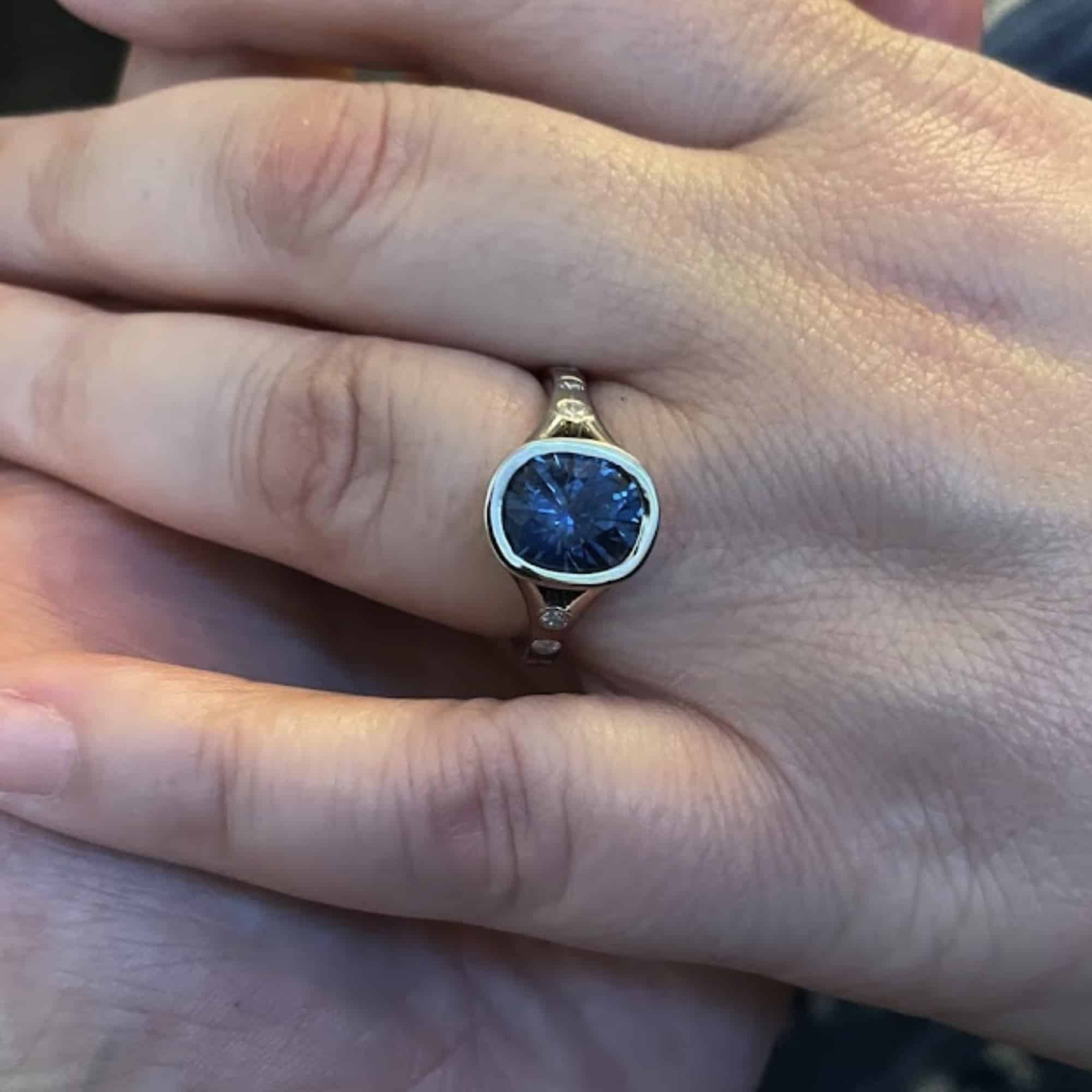 A photo from a customer review showing two people holding hands, one wearing a platinum "Corvus" ring with a large blue oval Montana sapphire
