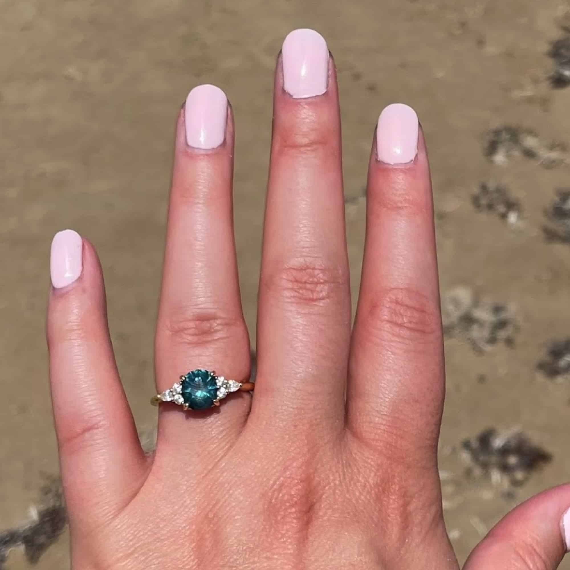 A photo from a customer review featuring a custom cluster ring with a large blue-green oval Montana sapphire on a baby-pink manicured hand against a beach
