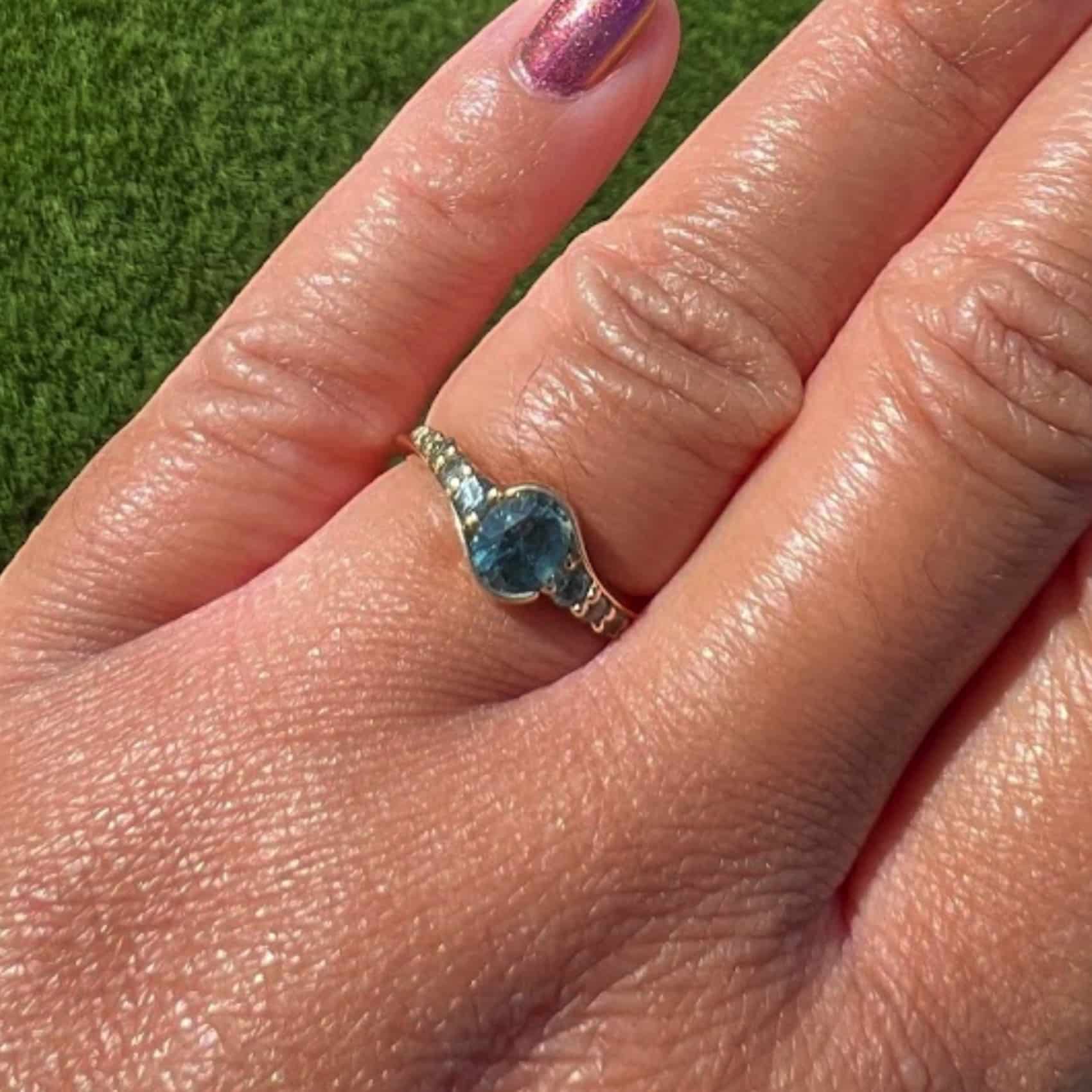 A photo from a customer review featuring a yellow gold "Venus" ring with a teal oval sapphire on a pink-manicured hand against grass