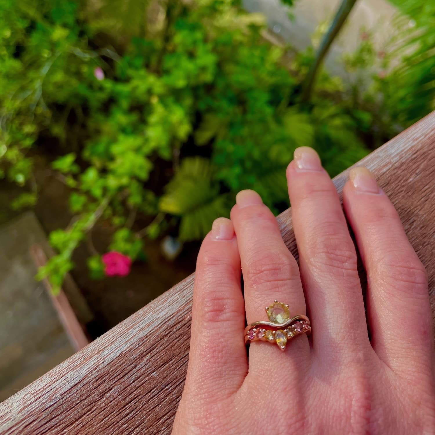 A photo from a customer review, featuring a yellow sapphire "Ruenna" ring and a custom sapphire "Merope" band on a well-manicured hand overlooking a garden.