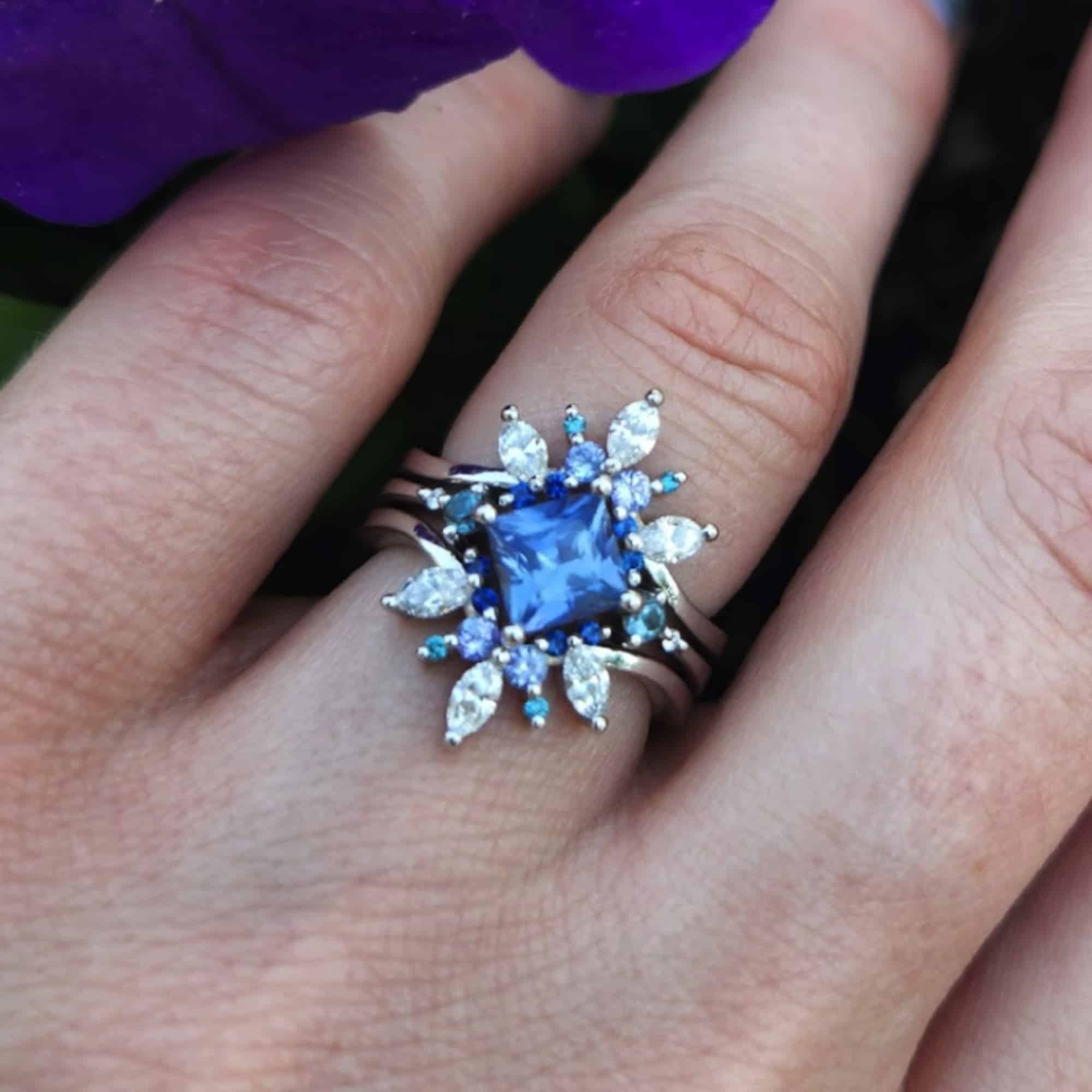 A photo from a customer review featuring a "Selene" ring with square blue sapphire and a "Selene" double band jacket on a hand against purple and pink flowers