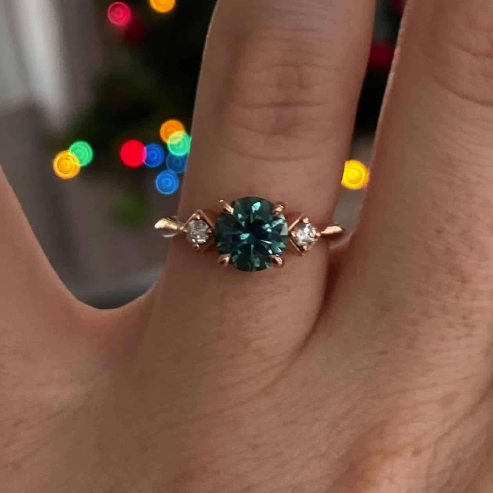 A photo from a customer review of a rose gold "Harmonia" ring with a green-teal round sapphire on a hand against twinkling Christmas lights.