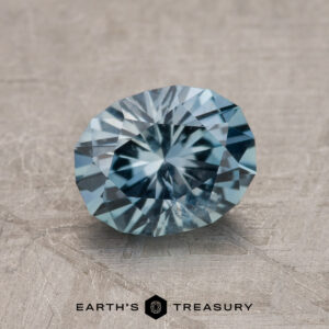 The 1.41-Carat Aqua Blue Montana Sapphire, with blue and green hues, is faceted in an oval shape and displayed on a textured gray surface. The "Earth's Treasury" logo and text are visible at the bottom.