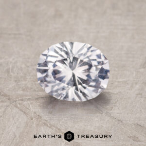 The 1.19-Carat Near Colorless Montana Sapphire, faceted in a brilliant oval cut, is displayed on a textured surface with "EARTH'S TREASURY" at the bottom.