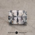 A 1.29-carat rectangular faceted Gray Montana Sapphire with a complex cut rests on a textured surface, featuring the "EARTH'S TREASURY" text at the bottom.