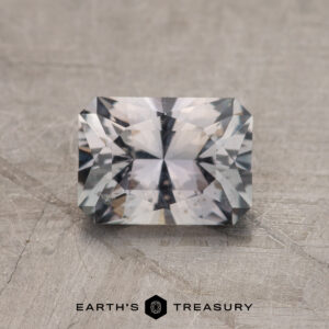 A 1.29-carat rectangular faceted Gray Montana Sapphire with a complex cut rests on a textured surface, featuring the "EARTH'S TREASURY" text at the bottom.