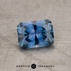 The 1.19-Carat Rich Steely Blue Montana Sapphire, with exquisite faceting and unique color, is showcased on a textured gray surface featuring the "EARTH'S TREASURY" logo at the bottom.