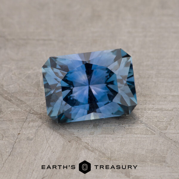 1.19-Carat Rich Steely Blue Montana Sapphire The 1.19-Carat Rich Steely Blue Montana Sapphire, with exquisite faceting and unique color, is showcased on a textured gray surface featuring the "EARTH'S TREASURY" logo at the bottom.