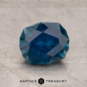 The 1.79-Carat Deep Peacock Blue Montana Sapphire (Heated) is shown upright on a textured gray surface with the Earth's Treasury logo and text beneath it.