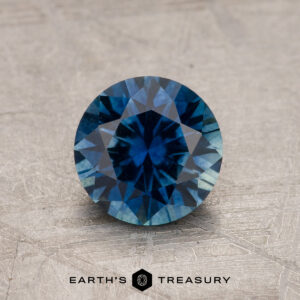 A 1.19-carat rich blue heated Montana Sapphire is displayed on a textured surface, with the "Earth's Treasury" logo and text beneath it.