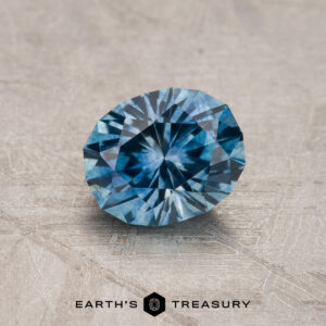 A 1.08-carat Teal Blue Montana Sapphire (heated) is shown on a textured gray surface, with "EARTH'S TREASURY" and two hexagonal logos beneath the gemstone.