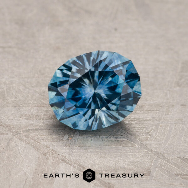 1.08-Carat Teal Blue Montana Sapphire (Heated) A 1.08-carat Teal Blue Montana Sapphire (heated) is shown on a textured gray surface, with "EARTH'S TREASURY" and two hexagonal logos beneath the gemstone.