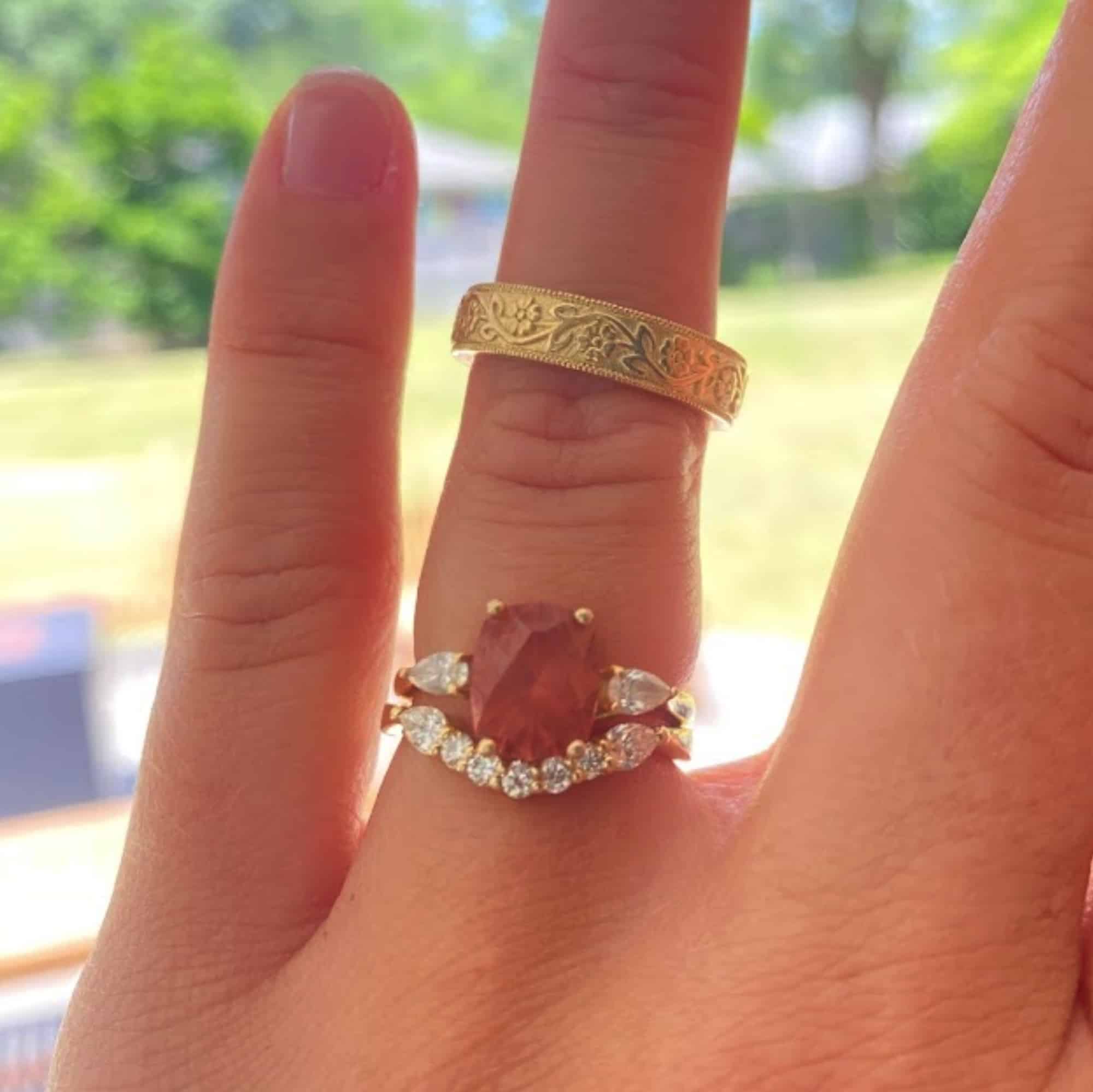 A photo from a customer review featuring a gold "Clematis" ring with orangey red garnet and custom diamond band, as well as a custom floral scroll men's band