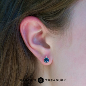 Close-up of a person's ear wearing Peacock Stella earrings, featuring a blue gemstone with a small clear stone above it. The "EARTH'S TREASURY" logo appears at the bottom of the image.