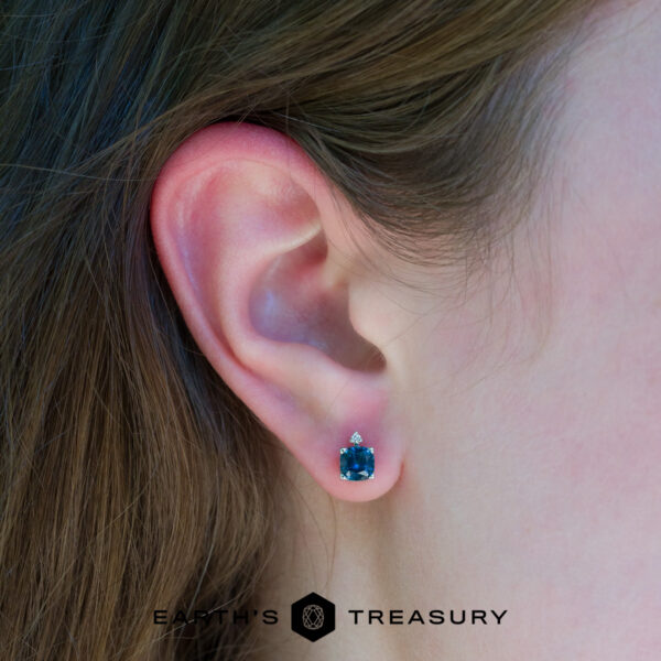 peacock stella Close-up of a person's ear wearing Peacock Stella earrings, featuring a blue gemstone with a small clear stone above it. The "EARTH'S TREASURY" logo appears at the bottom of the image.