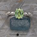 The "Olea” Hidden Halo Solitaire, a silver ring showcasing a green gemstone at its center and set with a twisted band design, is placed atop a blue rough gemstone. The background displays a textured gray stone surface adorned with geometric engravings. The text "EARTH'S TREASURY" is prominently visible at the bottom.