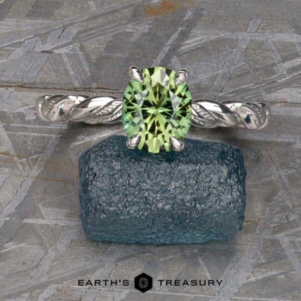 The Olea ring in platinum with 1.56-carat Australian sapphire The "Olea” Hidden Halo Solitaire, a silver ring showcasing a green gemstone at its center and set with a twisted band design, is placed atop a blue rough gemstone. The background displays a textured gray stone surface adorned with geometric engravings. The text "EARTH'S TREASURY" is prominently visible at the bottom.