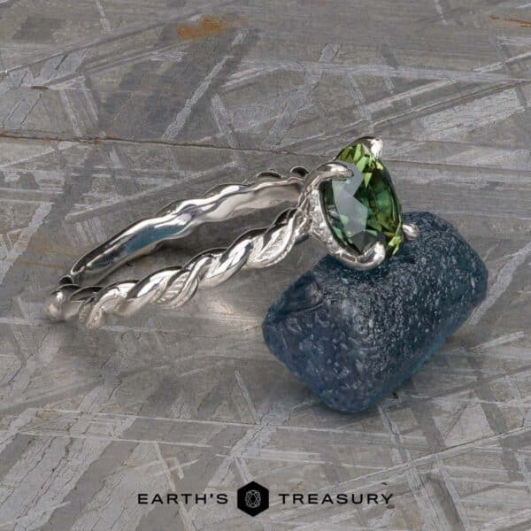 The Olea ring in platinum with 1.56-carat Australian sapphire The "Olea” Hidden Halo Solitaire, an elegant marquise-cut green gemstone ring with a twisted silver band, is showcased on a rough blue gemstone. The background has a textured, metallic appearance. At the bottom, the logo and text "EARTH'S TREASURY" are printed.