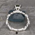 Close-up of the "Olea" Hidden Halo Solitaire ring featuring a green gemstone, seen from the side, showcasing its intricate braided band design. The ring is positioned upright on a textured blue rock against a gray background. The text "Earth's Treasury" is visible at the bottom of the image.