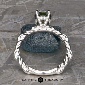 Close-up of the "Olea" Hidden Halo Solitaire ring featuring a green gemstone, seen from the side, showcasing its intricate braided band design. The ring is positioned upright on a textured blue rock against a gray background. The text "Earth's Treasury" is visible at the bottom of the image.