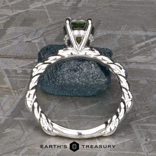 The Olea ring in platinum with 1.56-carat Australian sapphire Close-up of the "Olea" Hidden Halo Solitaire ring featuring a green gemstone, seen from the side, showcasing its intricate braided band design. The ring is positioned upright on a textured blue rock against a gray background. The text "Earth's Treasury" is visible at the bottom of the image.