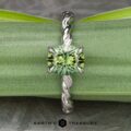 Displayed is "The 'Olea' Hidden Halo Solitaire," a stunning silver ring with an intricate, twisted band and a round, green gem at its center. The backdrop is filled with lush green leaves, and the text "EARTH'S TREASURY" is prominently featured at the bottom of the image.