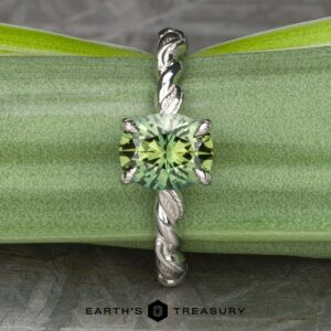Displayed is "The 'Olea' Hidden Halo Solitaire," a stunning silver ring with an intricate, twisted band and a round, green gem at its center. The backdrop is filled with lush green leaves, and the text "EARTH'S TREASURY" is prominently featured at the bottom of the image.